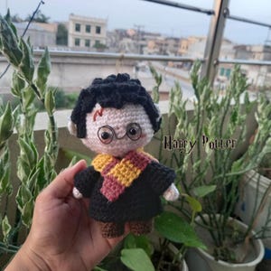 May include: A crocheted doll featuring black hair, round wire-rimmed glasses, and a red lightning bolt mark on the forehead. The doll wears a black robe with a yellow and red striped scarf. The text Harry Potter is visible on the right side.