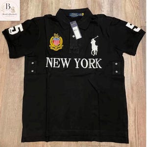 Puede incluir: Un polo negro de manga corta con el texto NEW YORK en blanco en el pecho. La prenda incluye un logotipo bordado de un jugador de polo en blanco, un escudo con emblema y el número 5 en ambas mangas. Cuenta con cuello y etiquetas.