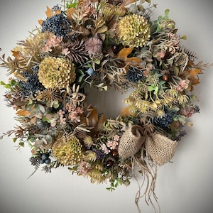 Pode incluir: Uma coroa circular composta por folhagem seca, incluindo folhas de carvalho castanhas, flores de hortênsia verdes, bagas azul-escuras e pequenas flores cor-de-rosa. Um laço de serapilheira está atado na base com fios de corda pendentes.