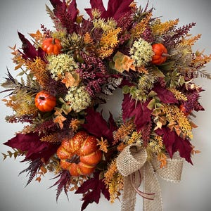 May include: A circular autumn wreath featuring deep burgundy maple leaves, orange pumpkins, yellow floral sprigs, and textured berry clusters. A natural tan burlap ribbon with two long hanging tails is tied at the bottom of the arrangement.
