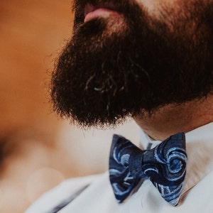 May include: A close-up of a man's beard and a blue bow tie with a swirling pattern. The bow tie is tied around the man's neck and is visible against a white shirt.