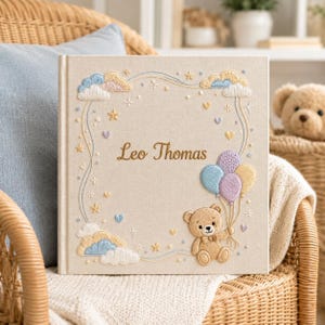May include: A square, beige fabric-covered photo album featuring embroidered details including clouds, stars, hearts, and a teddy bear holding three balloons. The name Leo Thomas is embroidered in the center in a brown script font.