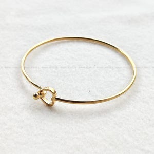 Può includere: Un bracciale rigido sottile color oro con un piccolo ciondolo a forma di cuore e una minuscola perlina sferica sulla chiusura. Il metallo ha una finitura lucida e riflettente. Il testo RAMA JEWELS è ripetuto in grigio chiaro nell'immagine.