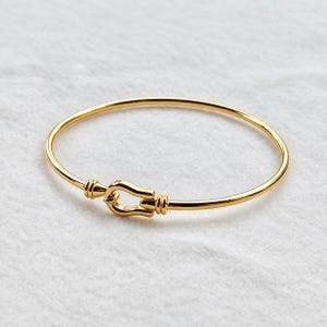 Pode incluir: Uma pulseira rígida de tom dourado polido com um mecanismo de fecho central em forma de ferradura. A banda circular é lisa e fina, ligando-se à peça em forma de U que é acentuada com pequenos detalhes cilíndricos estriados em cada lado.