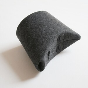 May include: A dark gray felt hat with a unique, angular design. The hat features a cylindrical base that tapers to a pointed end. Three oval-shaped cutouts are visible on the side. The hat is set against a white background.