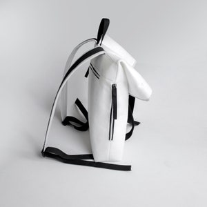 Puede incluir: Mochila blanca con correas negras y una cremallera. La mochila tiene una solapa que se pliega sobre la parte superior.