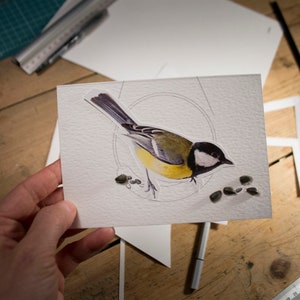 May include: A white card with a yellow and gray bird illustration and a circle drawn around it.  There are several small gray seeds scattered around the bird.