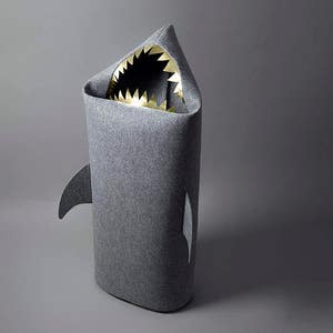 May include: A gray shark-shaped storage bin with a wide open mouth revealing gold-colored teeth. The bin has a fin on each side and is made of a soft, textured material. The bin is designed to look like a shark.