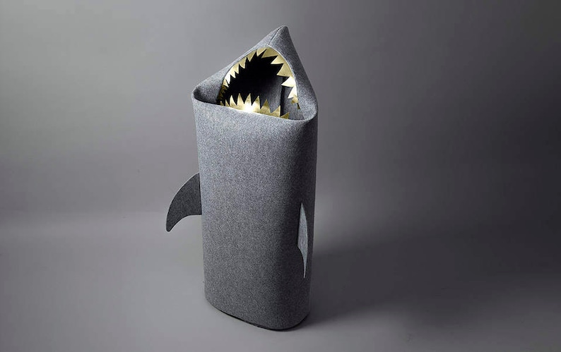 May include: A gray shark-shaped storage bin with a wide open mouth revealing gold-colored teeth. The bin has a fin on each side and is made of a soft, textured material. The bin is designed to look like a shark.