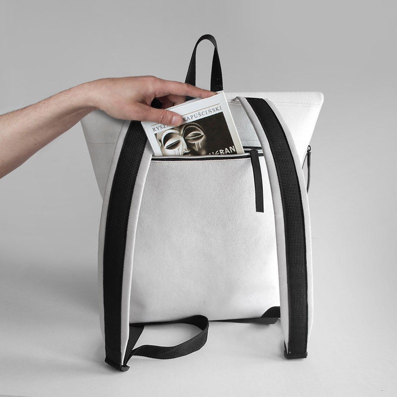 Puede incluir: Una mochila de lona blanca con correas negras. La mochila tiene un bolsillo con cremallera en la parte delantera con un libro titulado "Ryszard Kapuscinski -  The Shadow of the Sun" que sobresale.