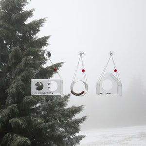 May include: Three clear acrylic bird feeders hanging from a window. Each feeder has a different shape, one is a rectangle with two holes, one is a circle, and one is a house shape. The feeders are suspended by thin strings with red beads at the top.