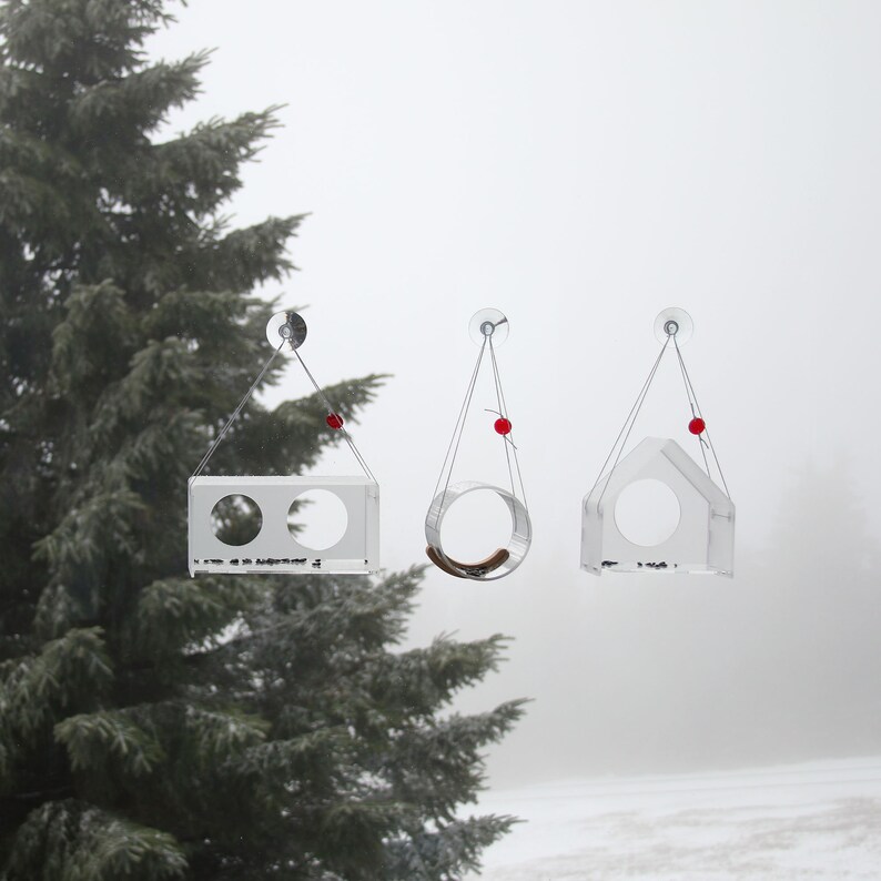 May include: Three clear acrylic bird feeders hanging from a window. Each feeder has a different shape, one is a rectangle with two holes, one is a circle, and one is a house shape. The feeders are suspended by thin strings with red beads at the top.