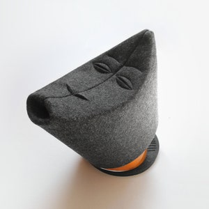 May include: A gray felt teapot with a stylized face design. The teapot has a triangular shape with a round base and a wooden accent. The design features embroidered eyes and a mouth.