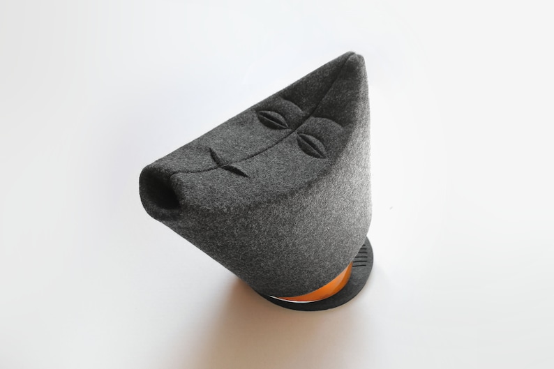 May include: A gray felt teapot with a stylized face design. The teapot has a triangular shape with a round base and a wooden accent. The design features embroidered eyes and a mouth.