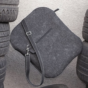 May include: A gray felt bag with a long adjustable strap. The bag has a zippered pocket on the front and is hanging against a stack of black tires.