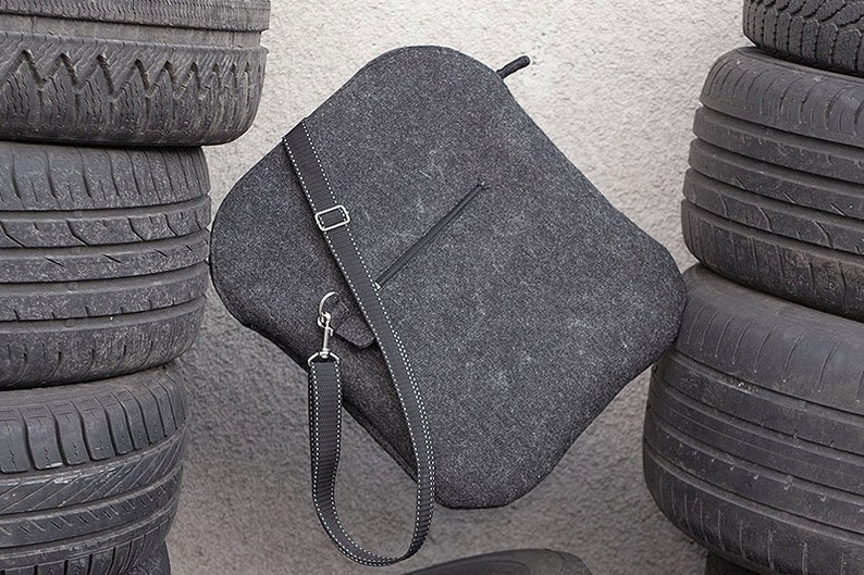 May include: A gray felt bag with a long adjustable strap. The bag has a zippered pocket on the front and is hanging against a stack of black tires.