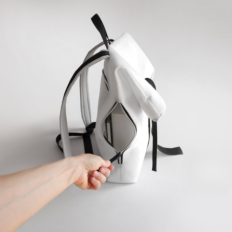Puede incluir: Una mochila blanca con correas negras y una cremallera. La mochila est&aacute; abierta y tiene un libro dentro.