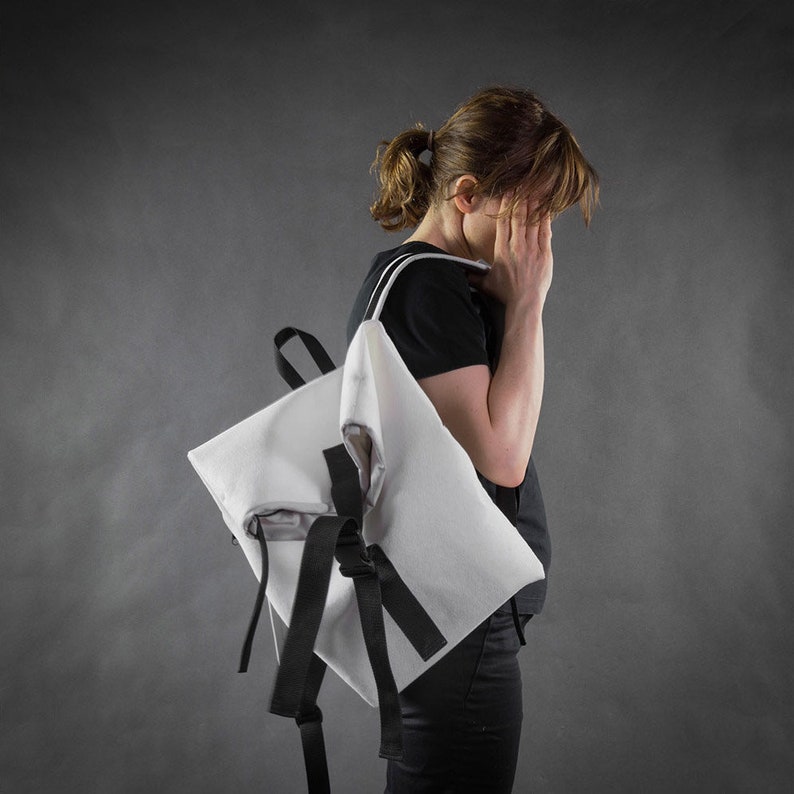 Puede incluir: Una mochila blanca con correas negras y una franja negra en la parte delantera. La mochila la lleva una persona con una coleta.