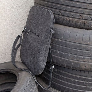 May include: A gray felt laptop sleeve with a zipper closure and a black adjustable shoulder strap. The sleeve is leaning against a stack of tires.