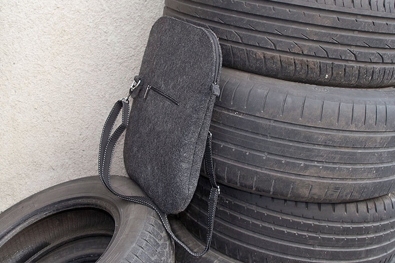 May include: A gray felt laptop sleeve with a zipper closure and a black adjustable shoulder strap. The sleeve is leaning against a stack of tires.