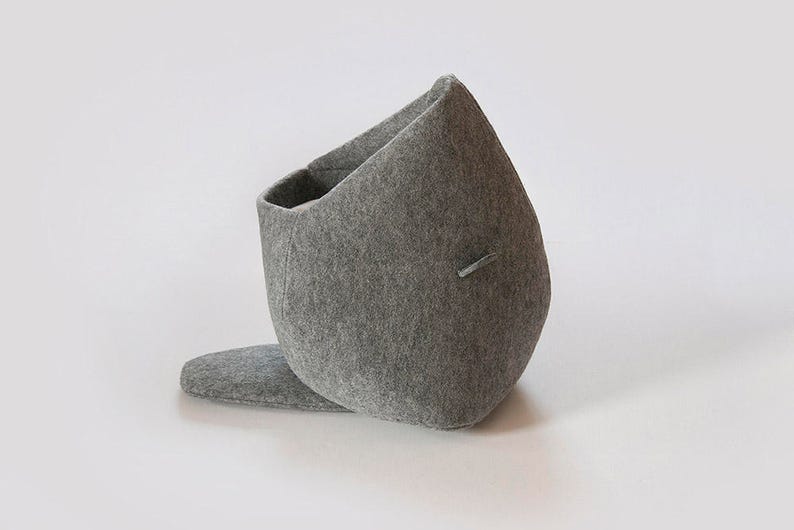 May include: A gray felt storage basket with a unique, asymmetrical design. The basket has a rounded body with a rectangular opening and a small, flat extension at the base. A small, rectangular detail is on the side.
