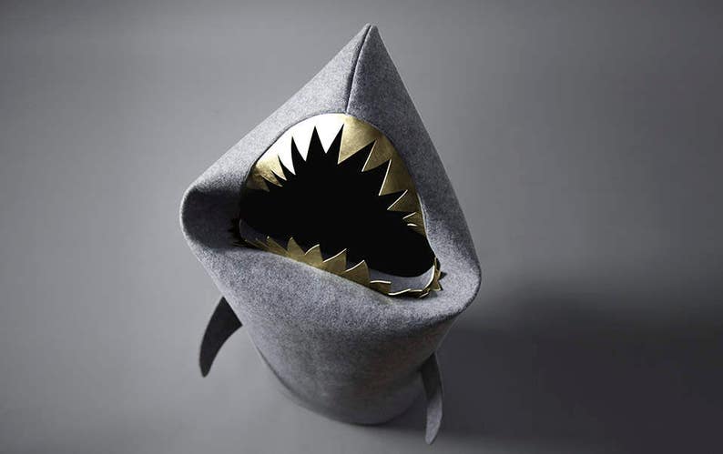 May include: A gray shark-shaped chair with a gold-colored mouth and teeth. The chair has a pointed fin on the top and two fins on the sides. The interior of the mouth is black.