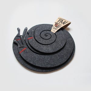Könnte beinhalten: Ein dunkelgrauer Filzuntersetzer in Form einer Schnecke mit einer roten Ziernaht am Kopf. Ein dreieckiges Pappschild ist oben befestigt und zeigt eine Grafik von gestapelten Pfannen sowie den Text PAN SNAIL in schwarzer und oranger Farbe.