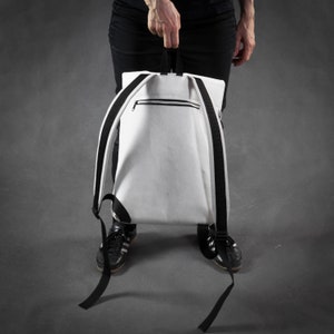 Puede incluir: Una mochila de lona blanca con correas negras y un bolsillo con cremallera en la parte delantera. La mochila la lleva una persona que lleva pantalones negros y zapatos negros.
