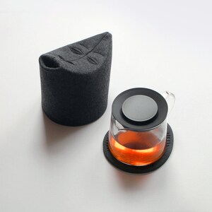 May include: A glass teapot with a black lid and a dark gray tea cozy. The teapot sits on a black coaster, and the tea inside is amber-colored. The cozy has a leaf design.