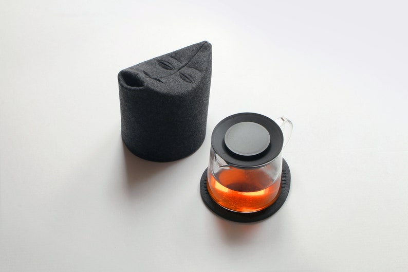 May include: A glass teapot with a black lid and a dark gray tea cozy. The teapot sits on a black coaster, and the tea inside is amber-colored. The cozy has a leaf design.