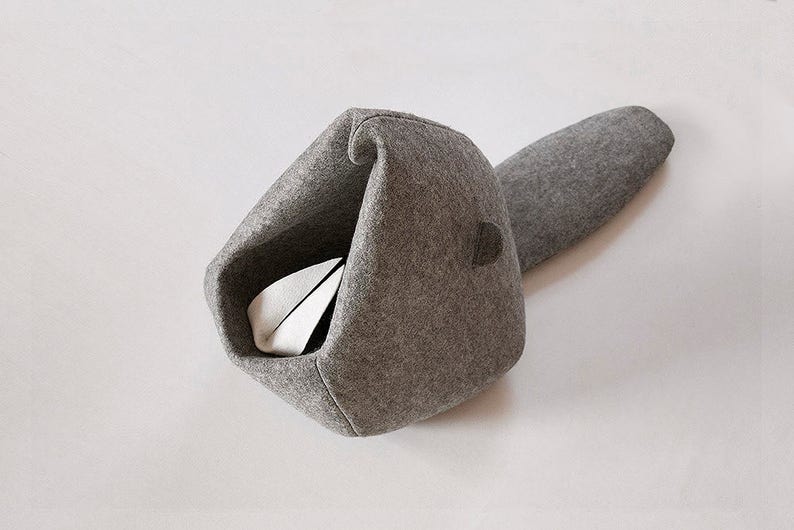 May include: A gray felt cat bed with a unique design. The bed has a rounded opening and a long, curved tail-like extension. Inside the bed is a white and black object. The bed is designed to provide a cozy and comfortable space for a cat to sleep or relax.