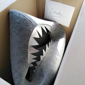 May include: A gray shark-shaped cushion with a white and black mouth, inside a cardboard box. The cushion is made of a soft, felt-like material. A handwritten note that says "Louke" is taped to the inside of the box.