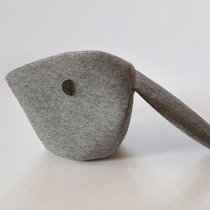 May include: A gray felt object with a rounded body and a long, straight extension. The body has a circular hole. The object is likely a decorative item or a unique piece of home decor. The felt material gives it a soft, textured appearance.