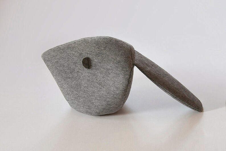 May include: A gray felt object with a rounded body and a long, straight extension. The body has a circular hole. The object is likely a decorative item or a unique piece of home decor. The felt material gives it a soft, textured appearance.