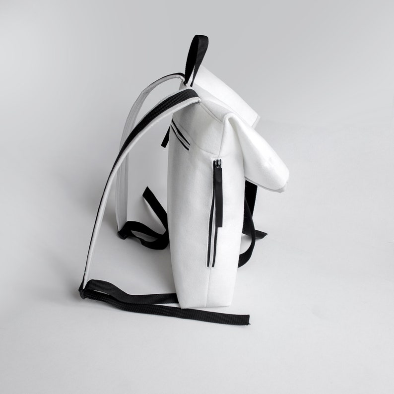 Puede incluir: Mochila blanca con correas negras y una cremallera. La mochila tiene una solapa que se pliega sobre la parte superior.
