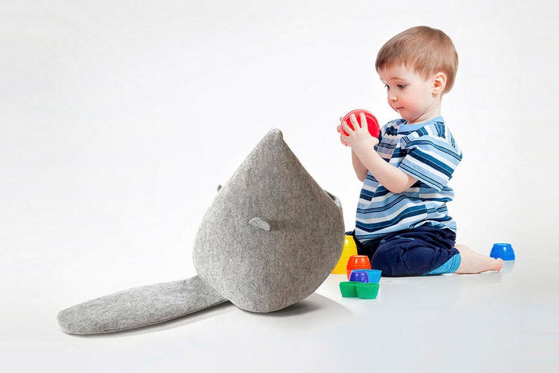 May include: A gray felt toy shaped like a whale with a long tail. A young child in a blue and white striped shirt and blue shorts is playing with a red ball. Colorful plastic cups and containers are scattered around.