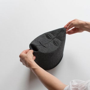 May include: A dark gray, leaf-shaped hat with a face design. The hat is made of a soft, felt-like material. The hat is being held up by two hands.