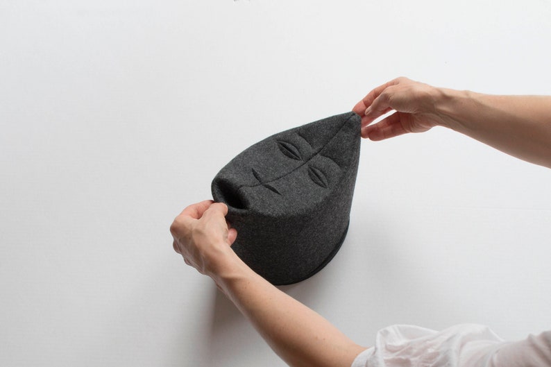 May include: A dark gray, leaf-shaped hat with a face design. The hat is made of a soft, felt-like material. The hat is being held up by two hands.