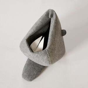 May include: A gray felt cat bed with a unique, modern design. The bed has a triangular shape with a deep interior, a small rounded detail on the side, and a long, curved base. Inside, a white object is visible.