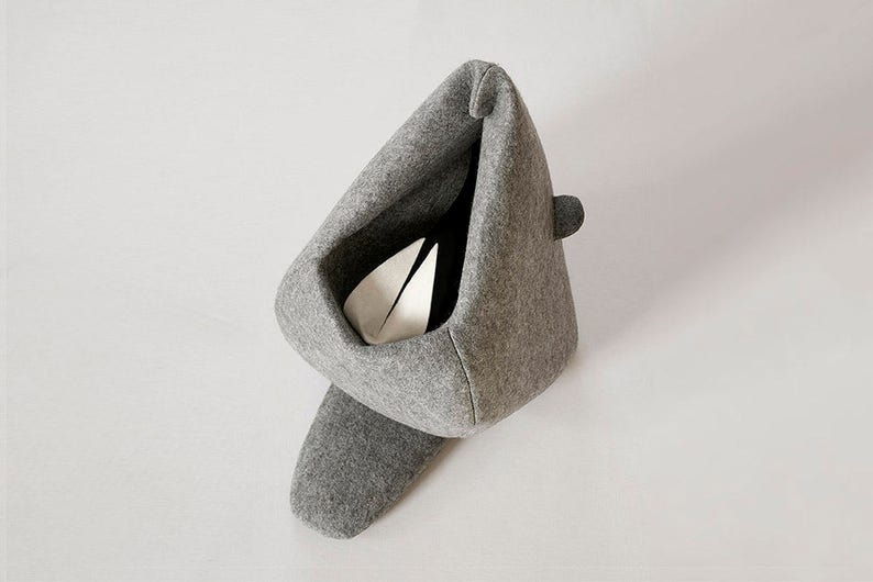 May include: A gray felt cat bed with a unique, modern design. The bed has a triangular shape with a deep interior, a small rounded detail on the side, and a long, curved base. Inside, a white object is visible.
