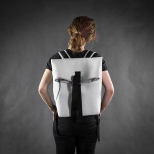 Puede incluir: Una persona con una camisa negra y una mochila blanca con una correa negra. La mochila tiene una solapa que se pliega sobre la parte superior.