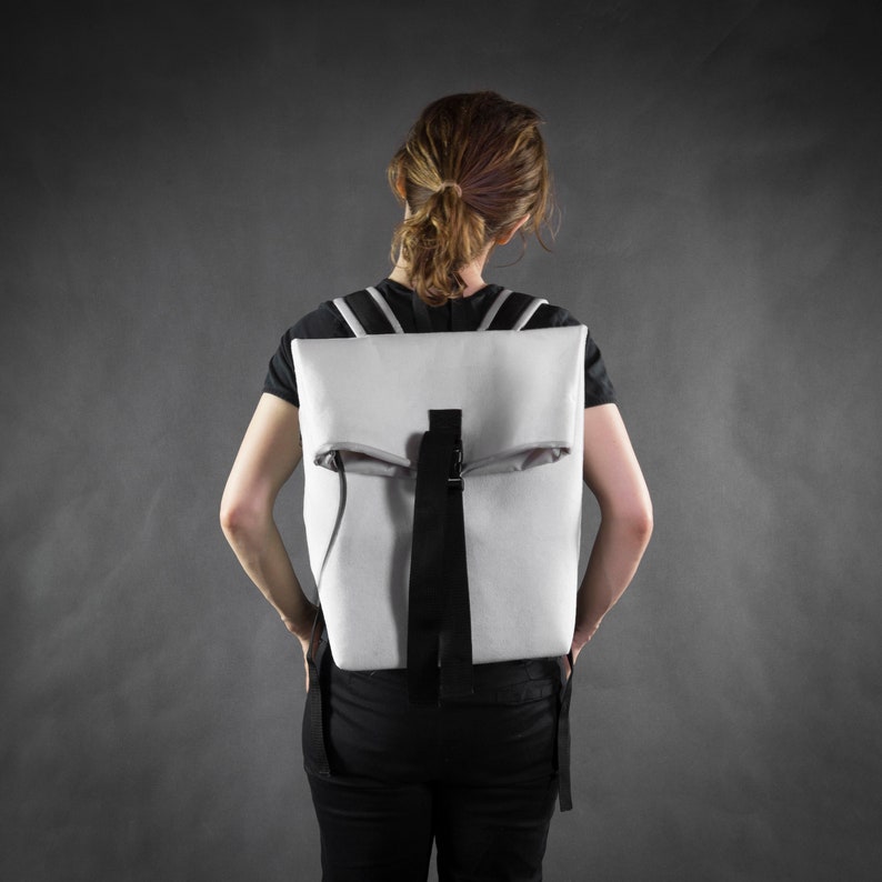 Puede incluir: Una persona con una camisa negra y una mochila blanca con una correa negra. La mochila tiene una solapa que se pliega sobre la parte superior.