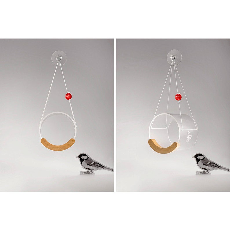May include: A clear acrylic bird feeder with a wooden perch hangs from a suction cup. A small bird is perched on the perch. The feeder has a red ball hanging from it.