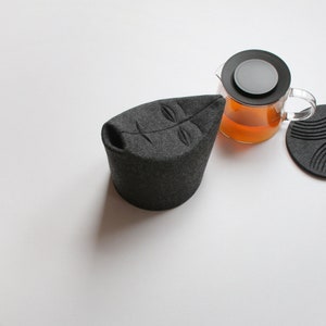 May include: A dark gray tea cozy shaped like a leaf, with a clear glass teapot filled with tea. Several dark gray coasters with various designs are also present, suggesting a tea set.