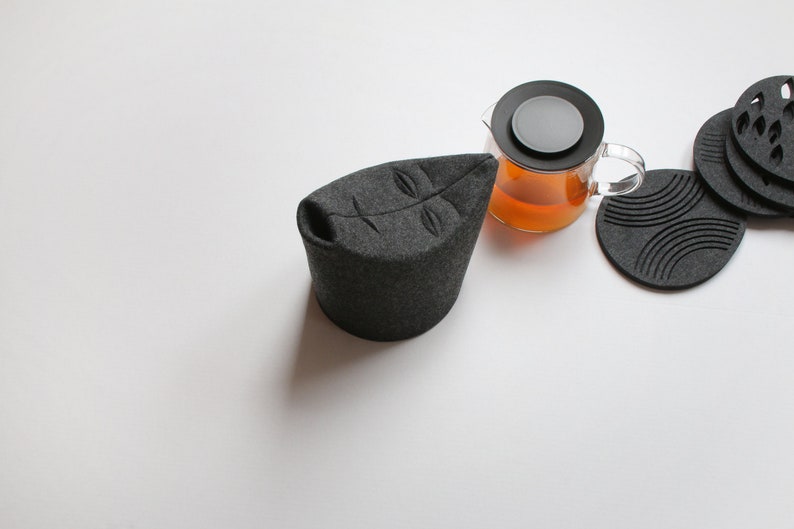 May include: A dark gray tea cozy shaped like a leaf, with a clear glass teapot filled with tea. Several dark gray coasters with various designs are also present, suggesting a tea set.