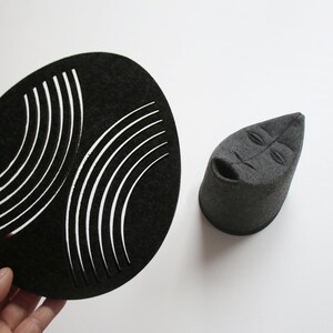May include: A black, oval-shaped felt piece with curved white cutouts, and a gray felt sculpture of a face with a cylindrical base. The objects are displayed on a white background.