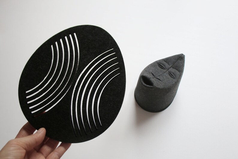 May include: A black, oval-shaped felt piece with curved white cutouts, and a gray felt sculpture of a face with a cylindrical base. The objects are displayed on a white background.
