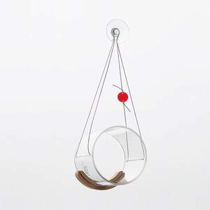 May include: A clear acrylic bird feeder with a wooden swing, hanging from a suction cup on a white background. A red bead is attached to the string.