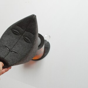 May include: A gray felt tea cozy in the shape of a leaf with embroidered facial features. The cozy is placed over a glass teapot with tea inside, sitting on a black coaster. The background is white.