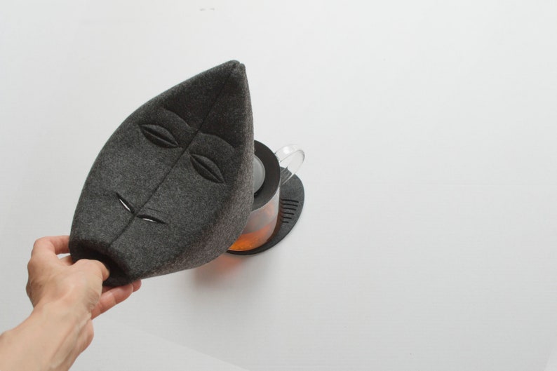 May include: A gray felt tea cozy in the shape of a leaf with embroidered facial features. The cozy is placed over a glass teapot with tea inside, sitting on a black coaster. The background is white.